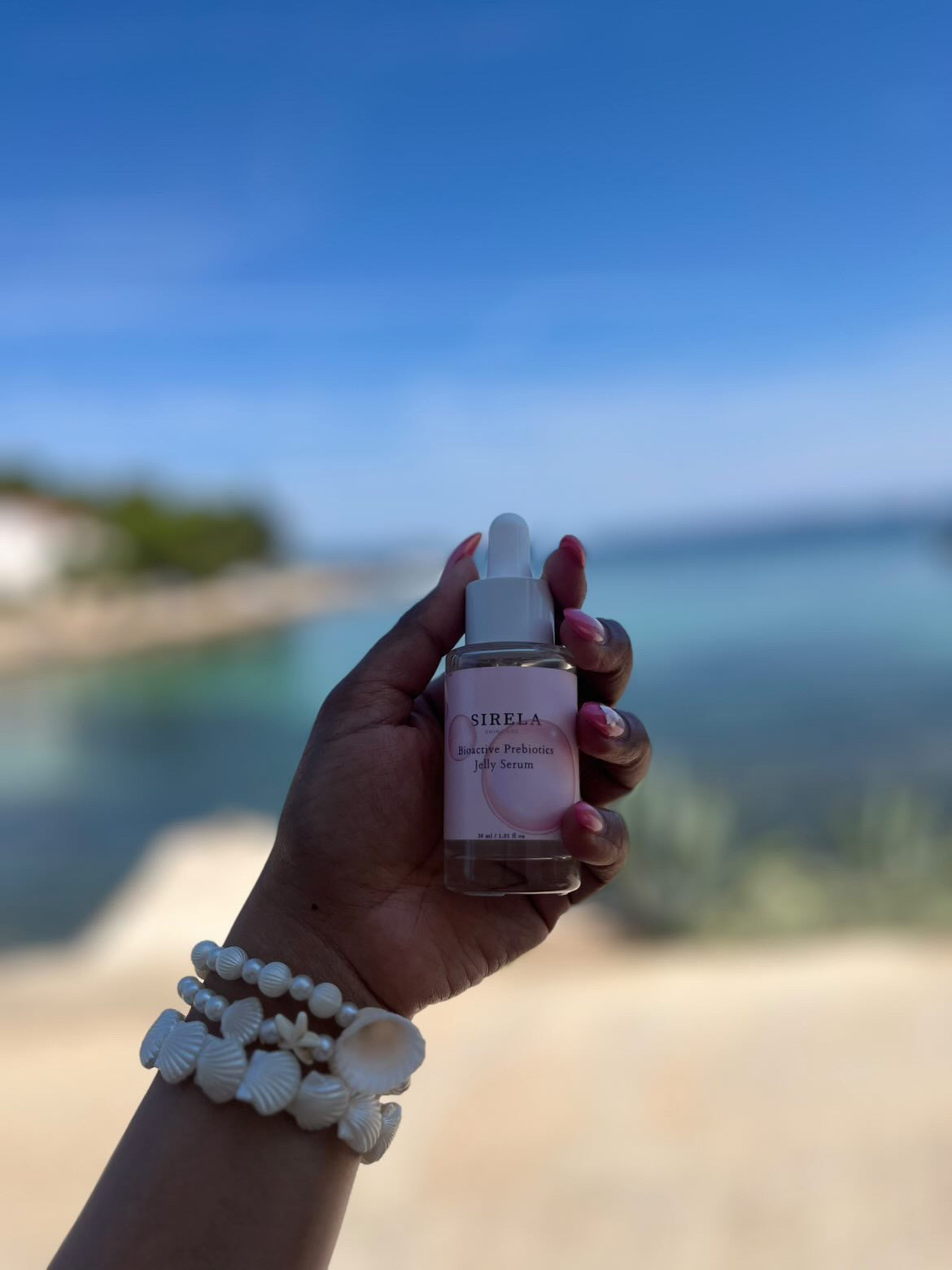 Hand holding a Sirela Skincare bottle with a pink label against a blurred beach background